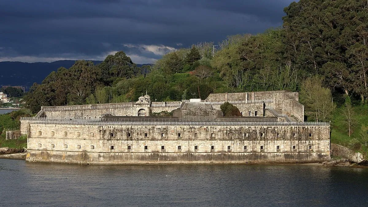 Muralla de piedra del castillo junto a la ría, con árboles y un cielo de tormenta