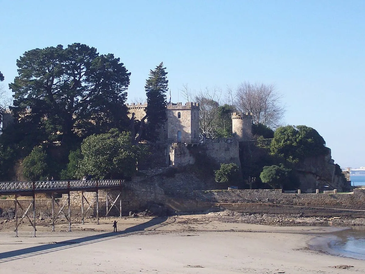 Castillo entre pinos sobre un islote, con la pasarela de madera que lo une a la playa