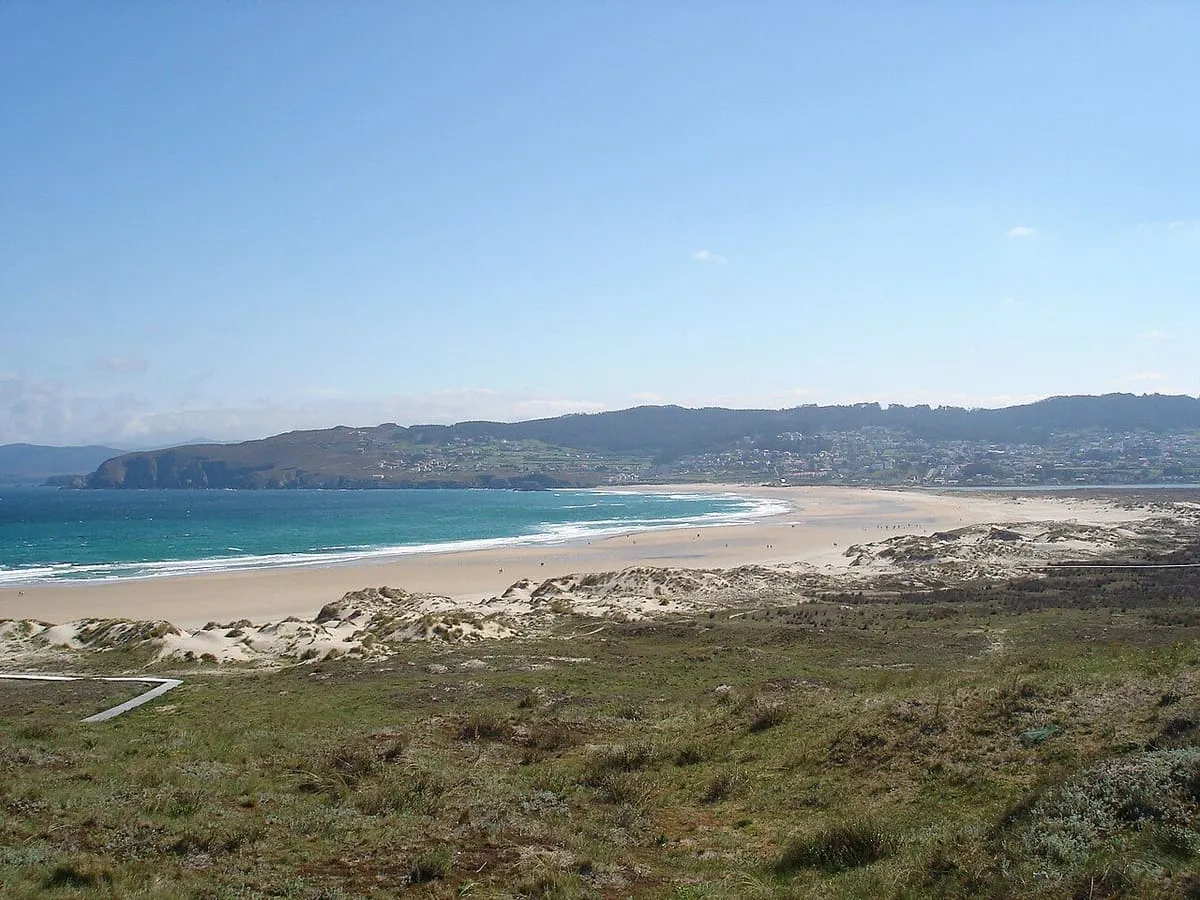 Playa larga de arena y dunas con una pasarela de madera, bajo un cielo despejado