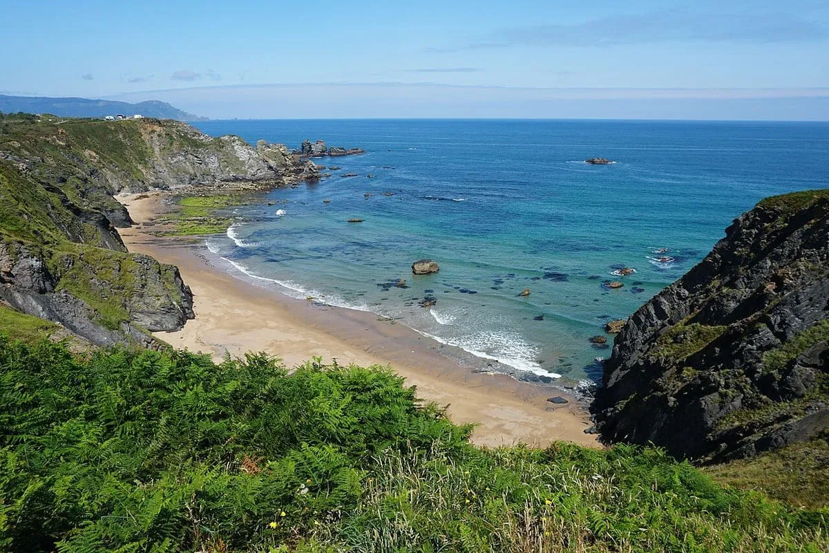 Playa de arena bajo acantilados cubiertos de hierba, con el mar turquesa