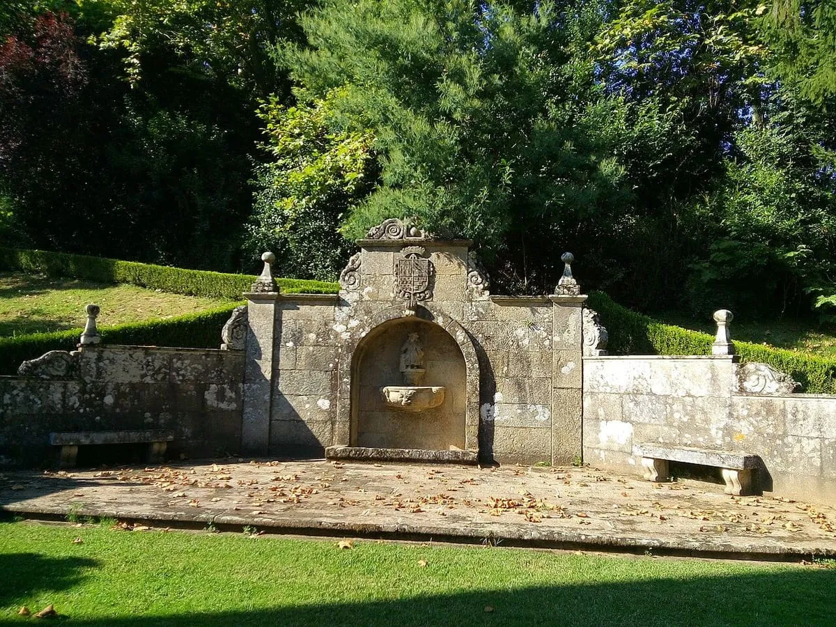 Fuente monumental de piedra con escudo y bancos, entre setos y árboles
