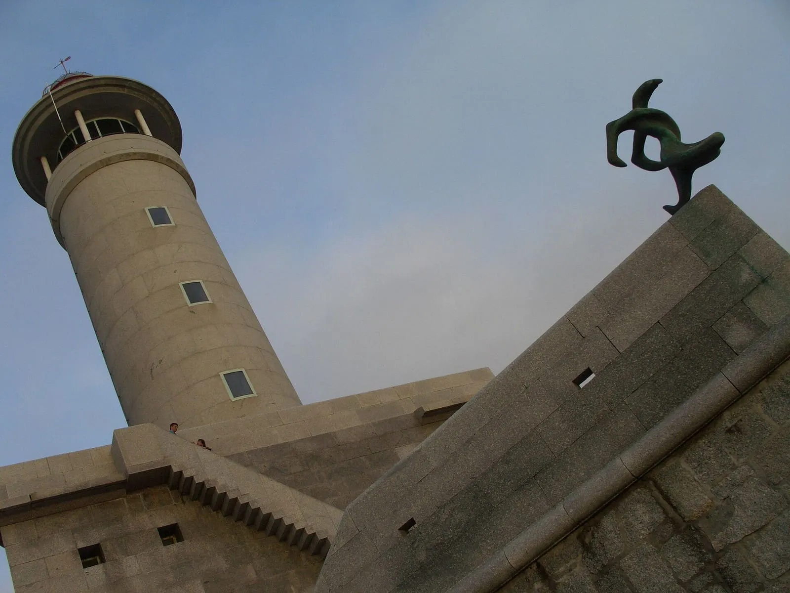 Torre del faro de Punta Nariga junto a una escultura, bajo un cielo nuboso
